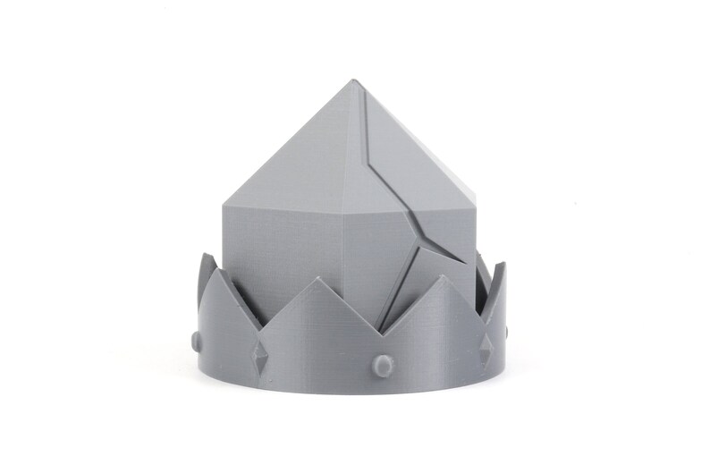 May include: A gray 3D printed crown with a pointed top and a jagged edge. The crown has a decorative design with a series of points around the base.