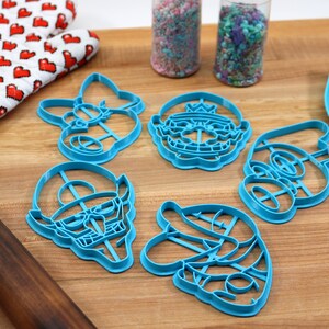 May include: A set of six blue plastic cookie cutters shaped like characters from a popular video game. The cutters are arranged on a wooden surface with a rolling pin, a spatula, and two jars of sprinkles.