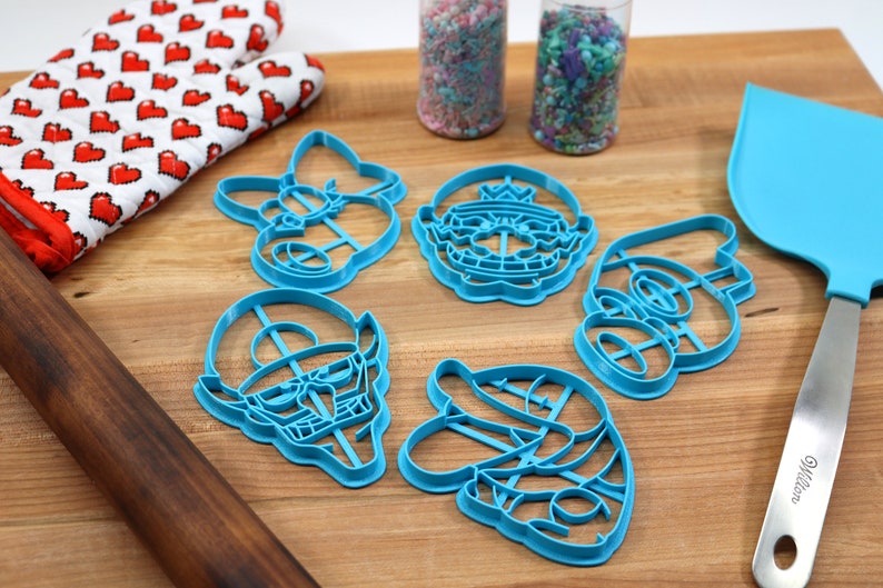 May include: A set of six blue plastic cookie cutters shaped like characters from a popular video game. The cutters are arranged on a wooden surface with a rolling pin, a spatula, and two jars of sprinkles.
