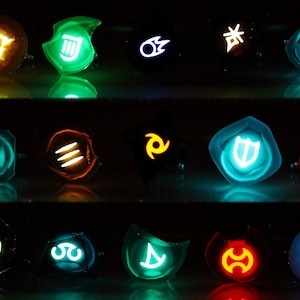 K&ouml;nnte beinhalten: Ein Set aus 15 leuchtenden Harzrringen mit verschiedenen Symbolen und Farben. Die Ringe sind in drei Reihen zu f&uuml;nf angeordnet. Die Symbole umfassen ein Schwert, einen Schild, ein Feuer, ein Wasser, einen Wind und eine Erde. Die Farben umfassen Blau, Gr&uuml;n, Rot, Gelb und Orange.