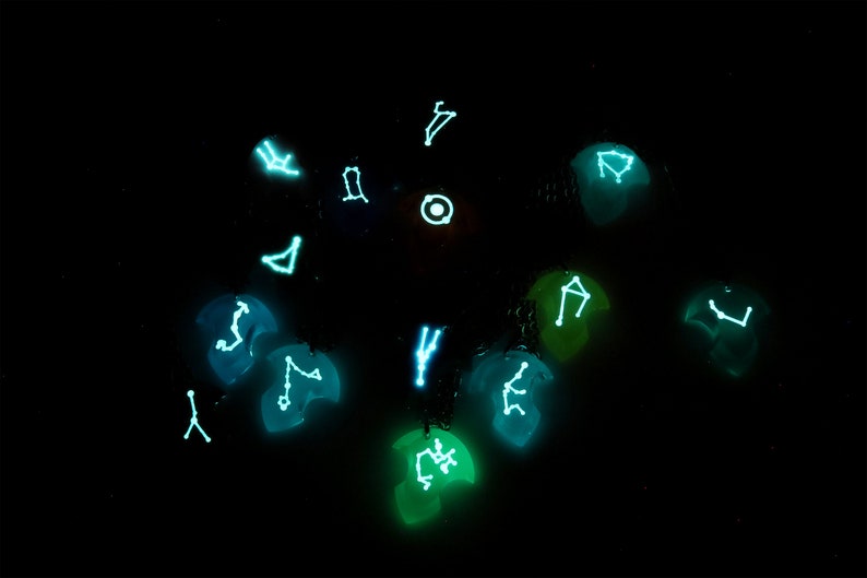 Puede incluir: Un conjunto de 12 dijes de constelaciones del zod&iacute;aco que brillan en la oscuridad. Cada dije es de un color diferente y presenta una constelaci&oacute;n diferente. Los dijes est&aacute;n dispuestos en c&iacute;rculo sobre un fondo negro.