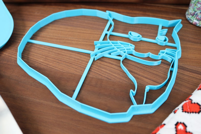 May include: A light blue plastic cookie cutter shaped like a cat's face. The cutter has a detailed design with a cat's eyes, nose, whiskers, and ears.