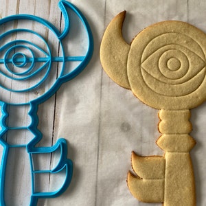 Op de afbeelding: Een blauwe plastic koekjesvormer in de vorm van een sleutel met een oogontwerp en een gele koek die in dezelfde vorm is gebakken.