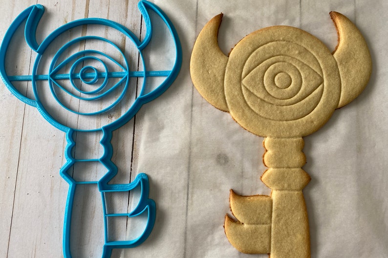 Op de afbeelding: Een blauwe plastic koekjesvormer in de vorm van een sleutel met een oogontwerp en een gele koek die in dezelfde vorm is gebakken.