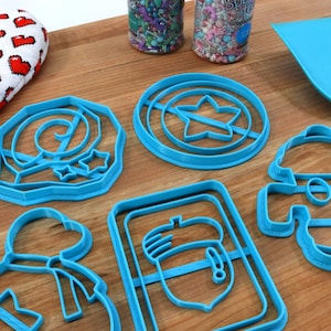 May include: A set of six blue plastic cookie cutters shaped like a snowman, a star, a mushroom, a bag with a bow, a swirl, and a shield with a star. The cutters are on a wooden surface.