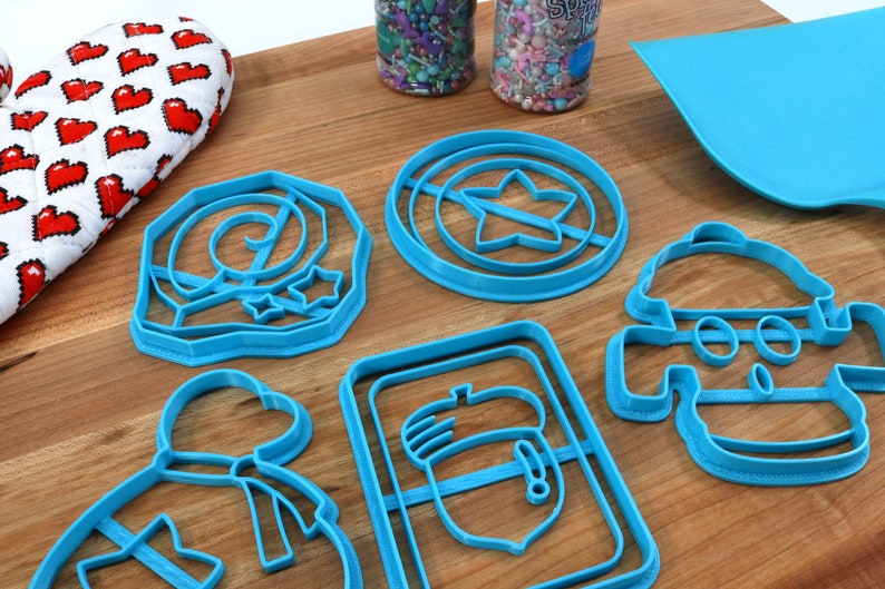 May include: A set of six blue plastic cookie cutters shaped like a snowman, a star, a mushroom, a bag with a bow, a swirl, and a shield with a star. The cutters are on a wooden surface.