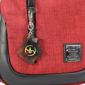 May include: A red crossbody bag with a black leather trim and a zipper closure. A brown and gold charm with a geometric design is attached to the zipper pull. The bag has a small silver label with the text "Made in Italy" and "Genuine Leather" on it.