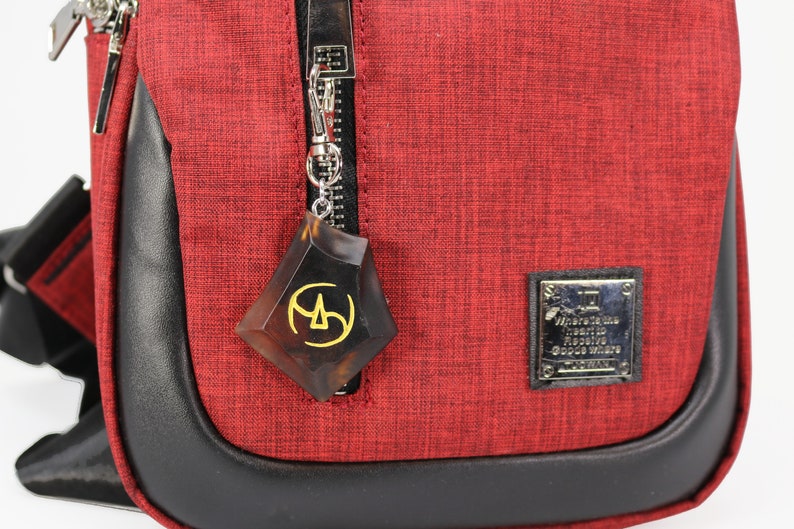 May include: A red crossbody bag with a black leather trim and a zipper closure. A brown and gold charm with a geometric design is attached to the zipper pull. The bag has a small silver label with the text "Made in Italy" and "Genuine Leather" on it.