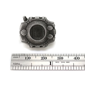 May include: A silver-colored metal pin shaped like a digital device with a screen and buttons. The pin is about 2 cm wide and is measured against a ruler.