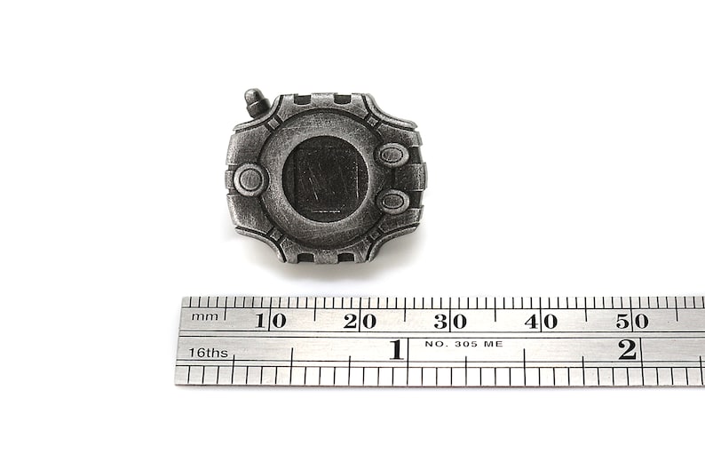 May include: A silver-colored metal pin shaped like a digital device with a screen and buttons. The pin is about 2 cm wide and is measured against a ruler.