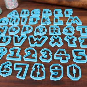May include: A set of blue plastic cookie cutters in the shape of the alphabet and numbers. The cutters are arranged on a wooden surface.