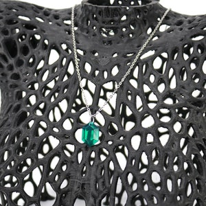 May include: A silver chain necklace with a dark green, faceted, geometric pendant. The necklace is displayed on a black, 3D printed, lattice-like mannequin.