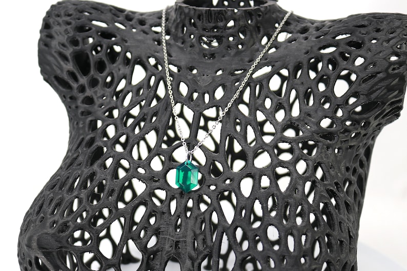 May include: A silver chain necklace with a dark green, faceted, geometric pendant. The necklace is displayed on a black, 3D printed, lattice-like mannequin.