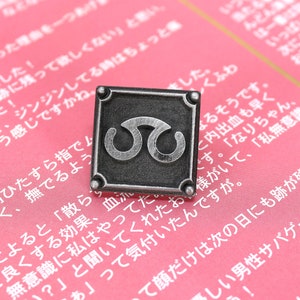 May include: A silver-toned metal pin with a square design. The pin features a stylized symbol of two interlocking circles. The pin is on a pink background.