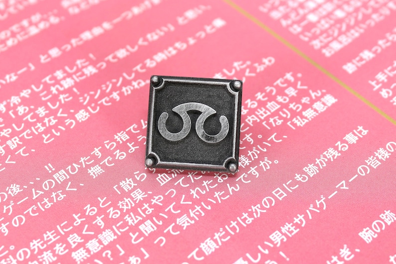 May include: A silver-toned metal pin with a square design. The pin features a stylized symbol of two interlocking circles. The pin is on a pink background.