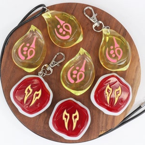 May include: A set of six resin charms featuring different designs. Three charms are yellow with pink symbols and come with a black cord necklace and a silver key chain. Three charms are red with gold symbols and come with a silver key chain.