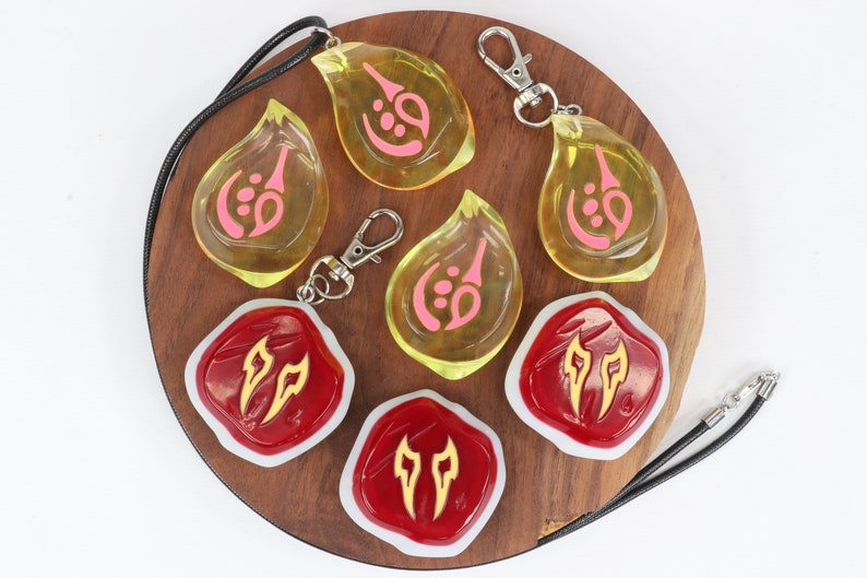 May include: A set of six resin charms featuring different designs. Three charms are yellow with pink symbols and come with a black cord necklace and a silver key chain. Three charms are red with gold symbols and come with a silver key chain.