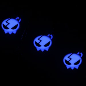 May include: Three glow-in-the-dark blue skull charms with a lightning bolt design. The charms are hanging from black cords.