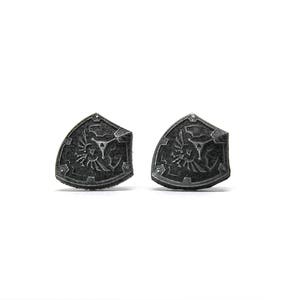 May include: A pair of silver-colored metal earrings shaped like a shield with a stylized design.