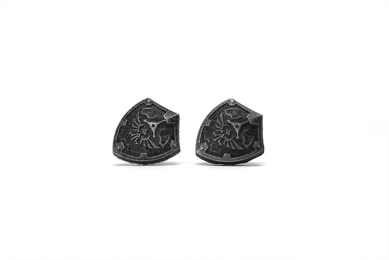 May include: A pair of silver-colored metal earrings shaped like a shield with a stylized design.