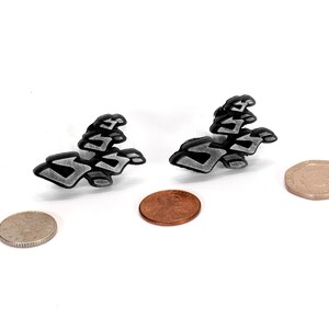 May include: Two silver-colored metal earrings shaped like abstract geometric designs. The earrings are shown on a white surface next to two coins.
