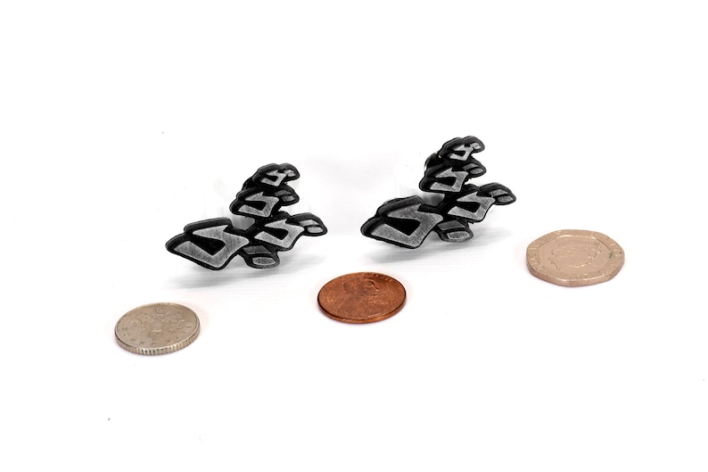 May include: Two silver-colored metal earrings shaped like abstract geometric designs. The earrings are shown on a white surface next to two coins.