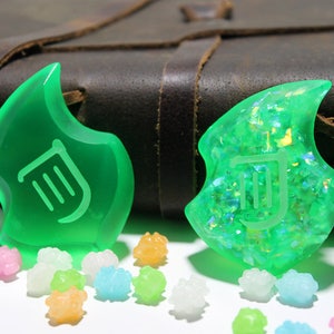 Op de afbeelding: Twee groene harscharms met een lier-symbool, een met een glinsterende afwerking, op een bruine lederen achtergrond met kleurrijke snoepjes.