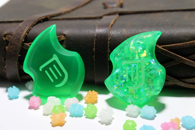 Op de afbeelding: Twee groene harscharms met een lier-symbool, een met een glinsterende afwerking, op een bruine lederen achtergrond met kleurrijke snoepjes.