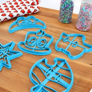 May include: A set of six blue plastic cookie cutters shaped like a hat, a snowman, a star, a ghost, a vest, and a cake. The cutters are on a wooden surface with sprinkles in jars and a rolling pin.