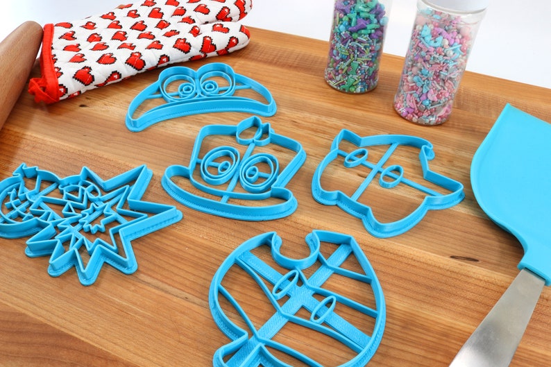 May include: A set of six blue plastic cookie cutters shaped like a hat, a snowman, a star, a ghost, a vest, and a cake. The cutters are on a wooden surface with sprinkles in jars and a rolling pin.