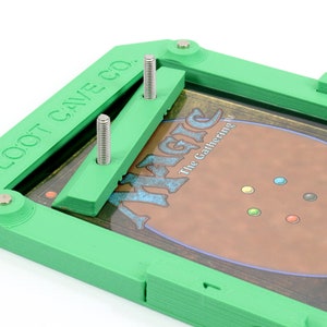 Op de afbeelding: Groene plastic kaarthouder met twee zilveren schroeven en de tekst "LOOT CAVE CO.". Een Magic: The Gathering kaart is zichtbaar in de houder.