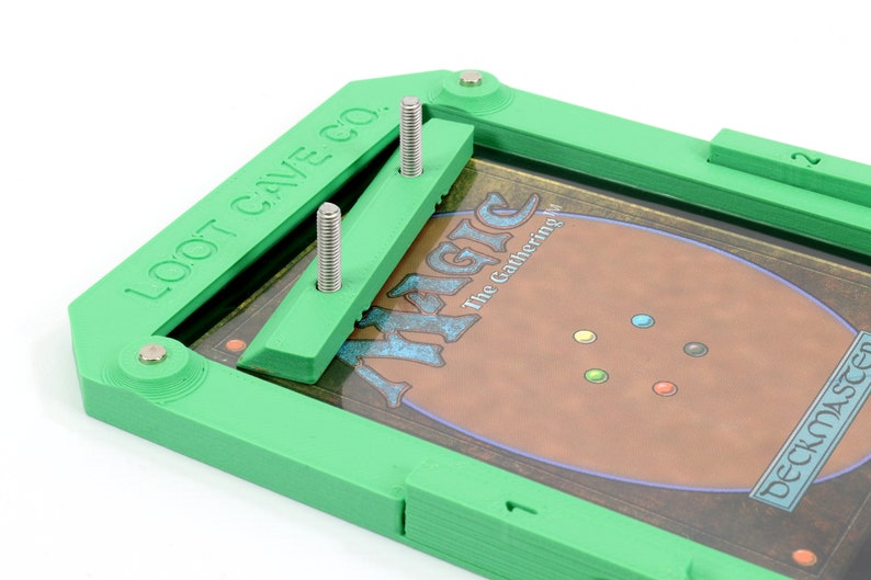Op de afbeelding: Groene plastic kaarthouder met twee zilveren schroeven en de tekst "LOOT CAVE CO.". Een Magic: The Gathering kaart is zichtbaar in de houder.