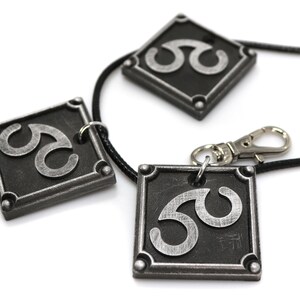 May include: Three silver-colored pendants with a black background and a stylized symbol on each. The pendants are attached to a black cord with a silver clasp.