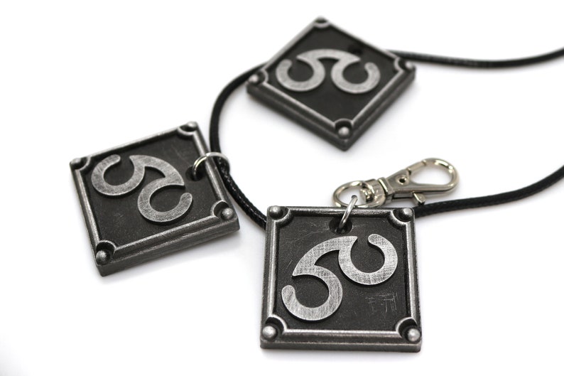 May include: Three silver-colored pendants with a black background and a stylized symbol on each. The pendants are attached to a black cord with a silver clasp.