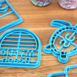 May include: A set of five blue plastic cookie cutters. The cutters are shaped like a palm tree, a crescent moon, a turtle, a beach umbrella, and a sign that says "My Little Beach".