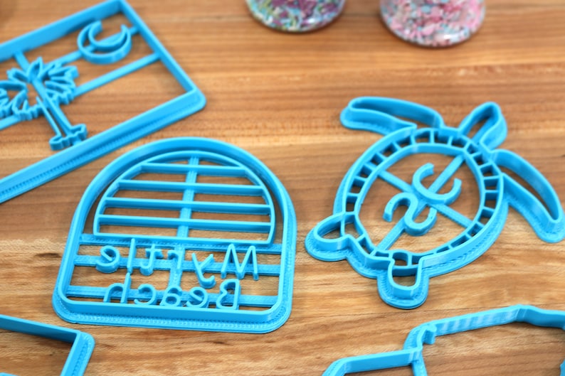 May include: A set of five blue plastic cookie cutters. The cutters are shaped like a palm tree, a crescent moon, a turtle, a beach umbrella, and a sign that says "My Little Beach".