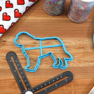 May include: A blue plastic dog-shaped cookie cutter, a blue spatula, and a black measuring tool are on a wooden surface. A red and white oven mitt with a heart pattern and two jars of sprinkles are also visible. Baking and crafting supplies.