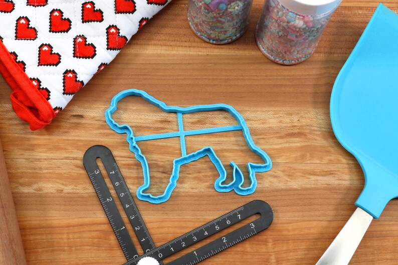 May include: A blue plastic dog-shaped cookie cutter, a blue spatula, and a black measuring tool are on a wooden surface. A red and white oven mitt with a heart pattern and two jars of sprinkles are also visible. Baking and crafting supplies.