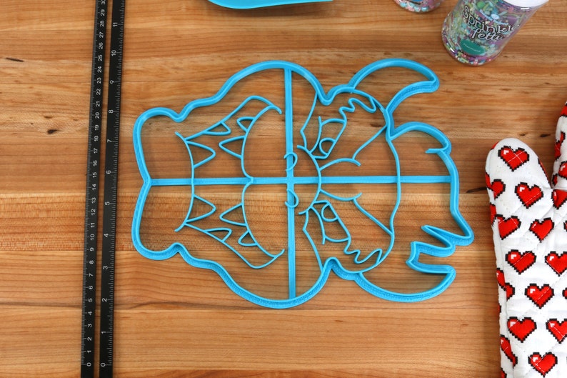 Op de afbeelding: Een blauwe plastic koekjesvormer in de vorm van een cartoonmonster met een brede grijns. De vormer is verdeeld in vier secties.