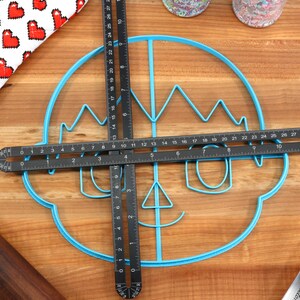 May include: A blue plastic cookie cutter shaped like a cartoon face with a crown, glasses, and a smile. Two black measuring tools are placed on top of the cookie cutter.