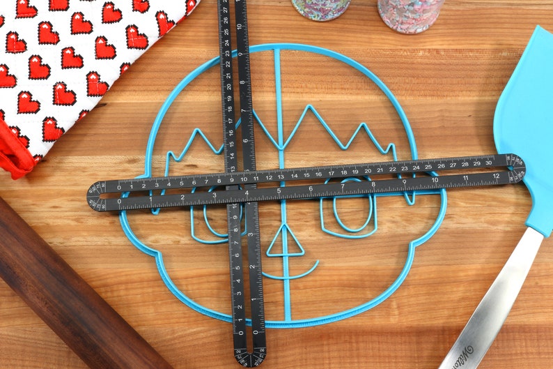 May include: A blue plastic cookie cutter shaped like a cartoon face with a crown, glasses, and a smile. Two black measuring tools are placed on top of the cookie cutter.