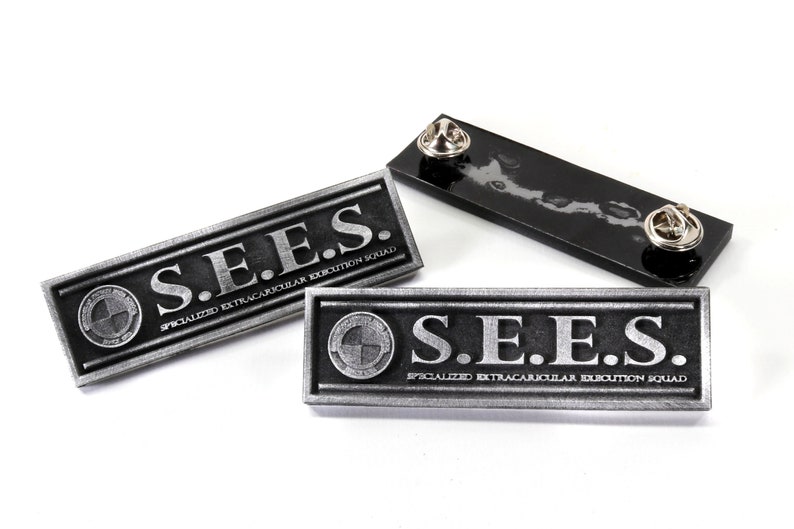 May include: Three silver metal pins with the text "S.E.E.S. SPECIALIZED EXTRACURRICULAR EXECUTION SQUAD" on them. The pins are rectangular and have a black background.
