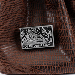 May include: A silver metal pin with a black and white image of a tree branch and the text "You are gunna carry that weight".