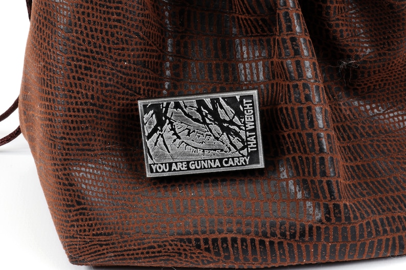 May include: A silver metal pin with a black and white image of a tree branch and the text "You are gunna carry that weight".