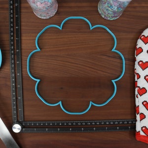 May include: A blue plastic cookie cutter shaped like a flower on a wooden surface. The cookie cutter is 15 cm in diameter.