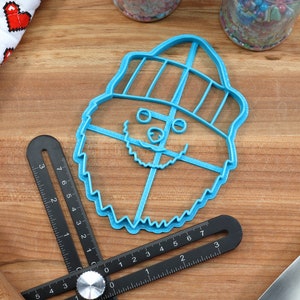 May include: A blue plastic cookie cutter shaped like a cartoon Yeti wearing a hat. The Yeti has a beard, eyes, and a nose.