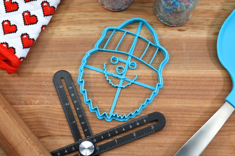 May include: A blue plastic cookie cutter shaped like a cartoon Yeti wearing a hat. The Yeti has a beard, eyes, and a nose.