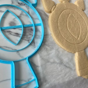 May include: A blue plastic cookie cutter shaped like a mirror with a handle, next to a light brown sugar cookie cut with the same shape.
