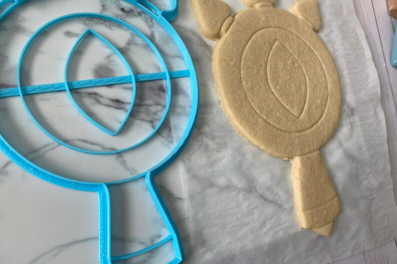 May include: A blue plastic cookie cutter shaped like a mirror with a handle, next to a light brown sugar cookie cut with the same shape.