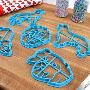 May include: A set of five blue plastic dog-shaped cookie cutters on a wooden surface. The cutters are in the shapes of a dog's head, a dog's body, and a dog's paw.